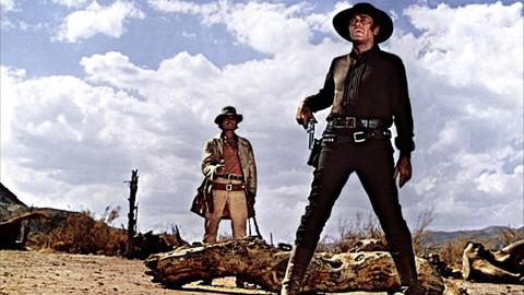 Das Bild zeigt eine Szene aus dem Italowestern "Spiel mir das Lied vom Tod" von 1968. Zu sehen sind zwei Männer mit Cowboy-Hüten, die in einer kargen Landschaft schräg hintereinander stehen. Beide haben Revolver gezogen.