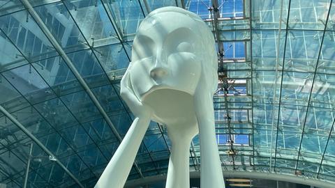 Das Bild zeigt eine weiße Skulptur am Fernbahnhof Frankfurt Flughafen. Die Figur besteht aus einem großen Kopf ohne Haare und mit geschlossenen Augen. Ihre Hände hat sie an ihre Ohren gelegt. Die Arme sind am Ellenbogen abgeknickt und ergeben eine Art Brücke, unter der Reisende hindurchgehen können.