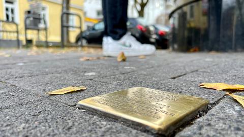 Nahaufnahme: Messingplatte im Boden mit unscharfer Schrift, im Hintergrund die Schuhe eines Passanten