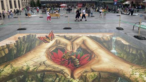 Das Bild zeigt Streetart in der Fuldaer Innenstadt. Zwei Hände formen ein Herz, in der Mitte ist eine Biene zu sehen, die auf einer roten Blüte sitzt. An der linken Hand sitzt an einem Fingerknöchel ein Schmetterling.