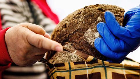 Eine Person mit blauer Einweghandschuhen überreicht einer anderen Person ein rundes, dunkles Laib Brot. Die empfangende Person hält eine Stofftasche mit einem Karomuster.