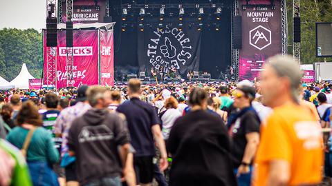 Eine Menschenmenge bewegt sich auf eine große Festivalbühne zu, die mit "Mountain Stage" beschriftet ist. Auf der Bühne sind Musiker unter einem Banner mit dem Text "TEAM SCHEISSE... GUTEN" zu sehen. Links im Bild steht ein Zelt mit der Aufschrift "Deutsches Rotes Kreuz" und Werbebanner von "Beck's". Rechts ist ein großer Bildschirm, der einen Gitarristen zeigt. Der Himmel ist bewölkt und Bäume sind im Hintergrund erkennbar.