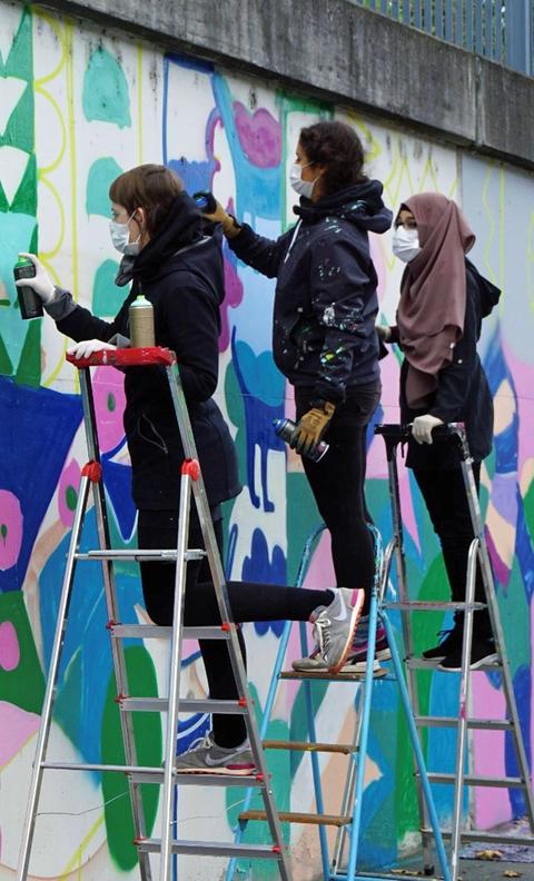 Drei junge Frauen stehen auf Leitern und besprühen eine Wand