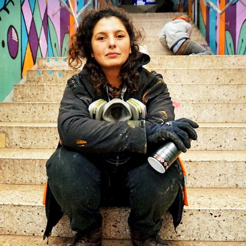 Eine junge Frau mit Atemmaske um den Hals und Spraydose in der Hand sitzt auf einer Treppe, die Wände sind bunt besprüht. Im Hintergrund kauert ein weiterer Mensch.