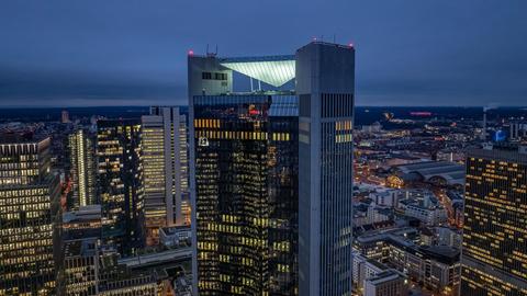 Die Abbildung zeigt eine nächtliche Skyline mit mehreren beleuchteten Hochhäusern. Im Vordergrund steht ein markantes Hochhaus mit einer Aussichtsplattform und einer pyramidenförmigen Struktur auf dem Dach. Die Fenster der Gebäude leuchten, und die Stadt breitet sich dahinter aus.