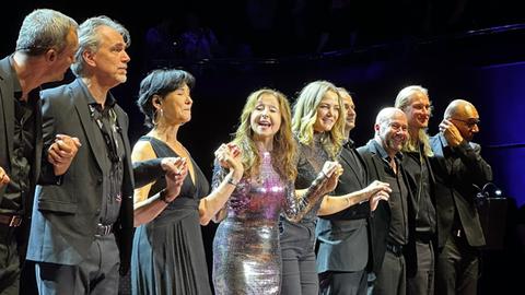 Das Bild zeigt Schlägersängerin Vicky Leandros mit ihrer Band auf einer Bühne. Sie trägt lange braune Haare mit Wellen und ein lila-glitzerndes Kleid und steht in der Mitte. Links und rechts von ihr stehen schwarz gekleidete Männer und Frauen. Alle halten sich an den Händen.