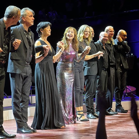 Das Bild zeigt Schlägersängerin Vicky Leandros mit ihrer Band auf einer Bühne. Sie trägt lange braune Haare mit Wellen und ein lila-glitzerndes Kleid und steht in der Mitte. Links und rechts von ihr stehen schwarz gekleidete Männer und Frauen. Alle halten sich an den Händen.