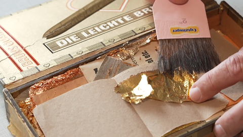 Eine Bürste und ein Finger bearbeiten Blattgold in einem Holzkästchen auf einem Tisch.