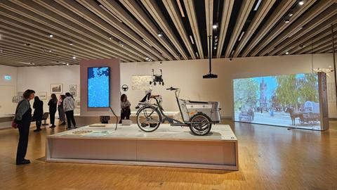 Das Bild zeigt einen Ausstellungsraum im Historischen Museum. Zu sehen ist ein großer rechteckiger Raum mit Holzboden, in dessen Mitte ein Podest steht. Darauf zu sehen ist ein Lastenfahrrad. An den Wänden sind verschiedene Bilder und eine Leinwand mit einer Videoinstallation zu sehen. Im Raum verteilt stehen Menschen, die sich Ausstellungsstücke ansehen.