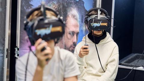 Das Bild zeigt zwei Schüler des Gymnasium Nord in Frankfurt, die das Spiel "Beethoven/Opus 360" ausprobiere. Sie sitzen hintereinander auf Stühlen und haben schwarze Virtual-Reality-Brillen vor den Augen. Sie tragen beide weiße Oberteile und halten Fernbedienungen in einer Hand, die mit einer Drahtspirale an der Brille festgemacht ist. 