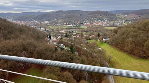 Im Bildvordergrund ein Handlauf aus Stahlseil. Dahinter eine Landschaft mit Ort im Tal und bewaldeten Hügeln.