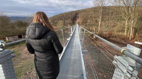 Im Bildvordergrund eine Frau von hinten, die in Richtung Brücker schaut. Dahinter eine sehr lange Fußgängerbücke, die über ein Tal zu einer bewaldeten Anhöhe führt.