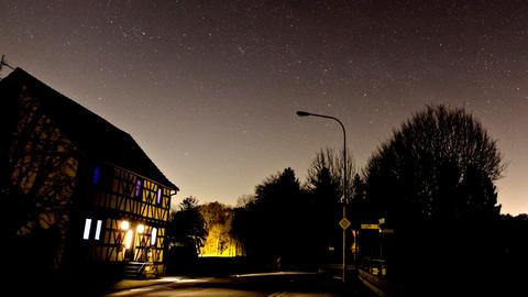 Foto, auf welchem eine nicht leuchtende Straßenlaterne zu sehen ist, die an einer Straße vor einem (leicht beleuchteten) Haus steht. Darüber ein Nachthimmel mit sehr vielen Sternen.