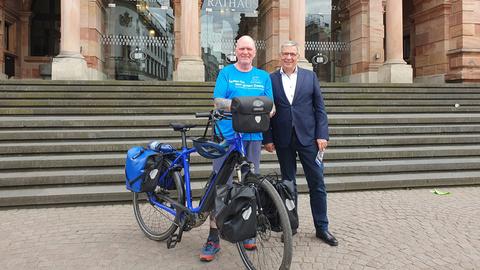 Oli steht mit seinem E-Bike vor dem Wiesbadener Rathaus. Neben ihm steht der Oberbürgermeister Gert-Uwe Mende. Beide lächeln freundlich. 