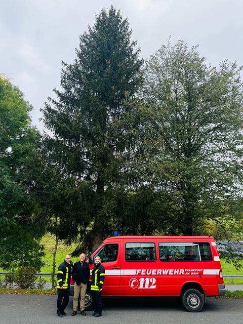 Drei Feuerwehrleute stehen vor einem Feuerwehrauto. Dhinter sind drei hochgewachsene Bäume zu sehen.