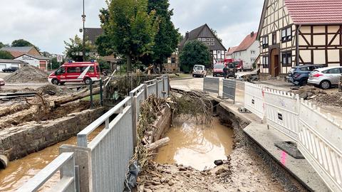 Foto von zerstörten baulichen Strukturen einer Kleinstadt: Eine großes Loch in der Straße, in dem braunes Wasser steht und ein abgebrochener Baum hineinragt. Daneben viele Absperrgitter und im Hintergrund Einsatzfahrzeuge der Feuerwehr.