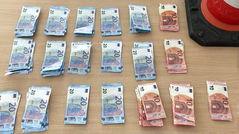 10- und 20-Euro-Noten liegen geordnet in Stapeln auf einem Tisch (von oben fotografiert). Daneben steht eine kegelförmige orangfarbene Verkehrsboje/-spitze der Polizei