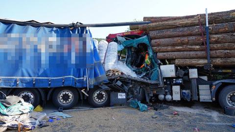 Das Bild zeigt zwei Lkw, die ineinandergefahren sind. Die Ladefläche des vorderen Lkw ist mit einer blauen Plane verkleidet. Die Fahrerkabine des hinteren, mit Baumstämmen beladenen Lkw ist zusammengedrückt.