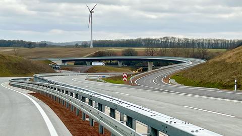 Foto von neugebauten Autobahnen. Der Blickwinkel ist so gewählt, dass sich die Straßen streifenartig, geschwungen ineinander "verweben". Obendrüber bzw. im Hintergrund grauer Himmel mit einem Windrad.