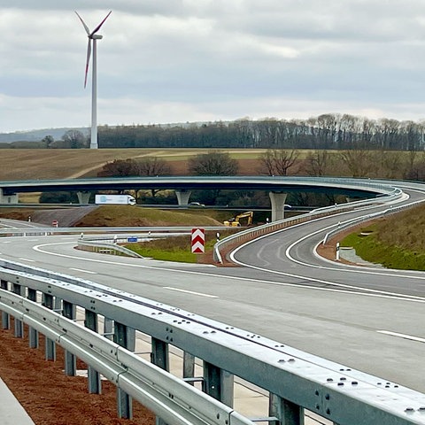 Foto von neugebauten Autobahnen. Der Blickwinkel ist so gewählt, dass sich die Straßen streifenartig, geschwungen ineinander "verweben". Obendrüber bzw. im Hintergrund grauer Himmel mit einem Windrad.