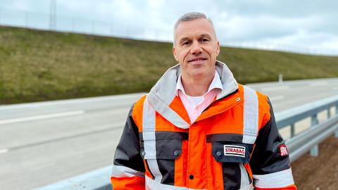 Foto eines Mannes in neonfarbener Arbeitskleidung mit einem Schild "STRABAG" an der Brust, der in die Kamera lächelt. Im Hintergrund unscharf eine leere Autobahn.