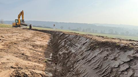 Ein gelber Bagger steht auf einem Feld. Im Vordergrund ist ein ausgehobener Graben zu sehen.