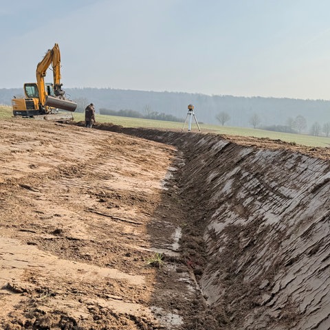 Ein gelber Bagger steht auf einem Feld. Im Vordergrund ist ein ausgehobener Graben zu sehen.