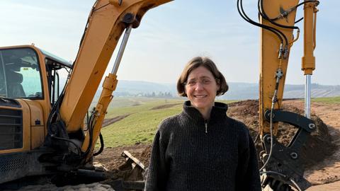 Eine Frau mit Brille lächelt in die Kamera. Im Hintergrund steht ein Bagger.