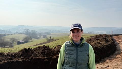 Eine Frau steht auf einem Feld. Sie trägt eine Cap und eine grüne Daunenweste.