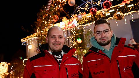 Dominik Piezcko (l.) und Sascha Bärwald in roten Winterjacken vor ihrem im Dunklen beleuchteten Haus, das mit tausenden Lichtern weihnachtlich geschmückt ist