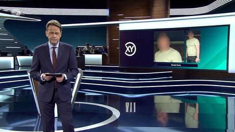 Fernsehstudio, vorner steht ein dunkelblonder Moderator im Anzug, auf einen Bildschirm ist ein kurzhaariger, etwas korpulenter Mann mit kurzen Haaren projiziert.