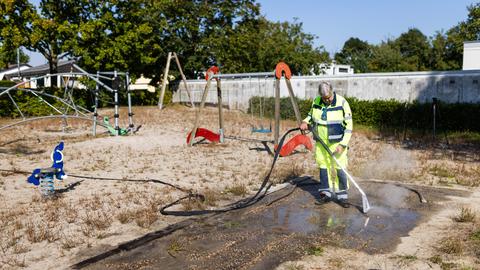 Ein Mann in Arbeitskleidung steht auf einem Spielplatz und hält einen Hochdruck-Wasserschlauch. 