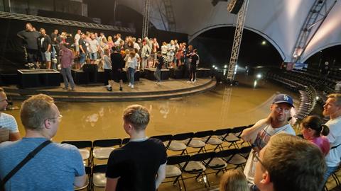 Eine Open-Air-Bühne unter einem Zeltdach steht unter Wasser. Zuschauer stehen auf Podesten und Stühlen.