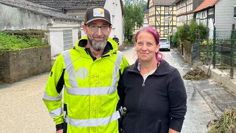 Ein Mann in Warnweste und eine Frau stehen auf einer leicht überschwemmten Straße.