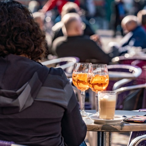 Besucher eines Straßencafes in der Innenstadt von Darmstadt im Sonnenschein, im Vordergrund eine Frau von hinten vor zwei Drinks und einem Kaffee