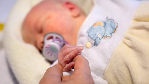 Ein schlafendes Baby liegt auf einer Decke und greift mit seinen Fingern nach einer Hand.