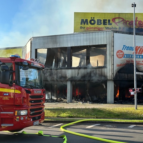 Ausgebranntes Fabrikgebäude, das ein Möbelhaus ist, davor Feuerwehrwagen, darüber Rauch