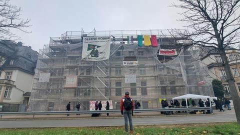 Eingerüstetes vierstöckiges Haus mit einem Banner mit der Aufschrift "Defend Rojava", davor ein Zelt mit mehreren Menschen