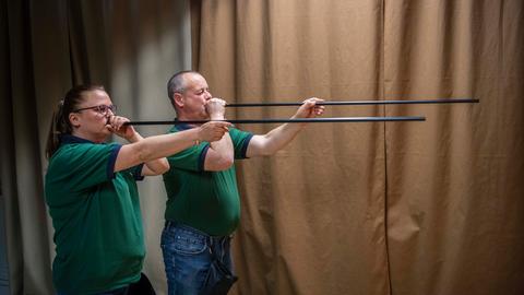 Zwei Personen stehen nebeneinander und halten Blasrohre an ihre Münder, als ob sie schießen. Beide tragen grüne Poloshirts und blaue Jeans. Der Hintergrund besteht aus einem beigen Vorhang.