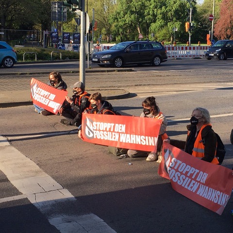 Sechs Menschen mit Warnwesten sitzen auf einer Straße. Sie halten Transparente mit der Aufschrift "Stoppt den fossilen Wahnsinn".