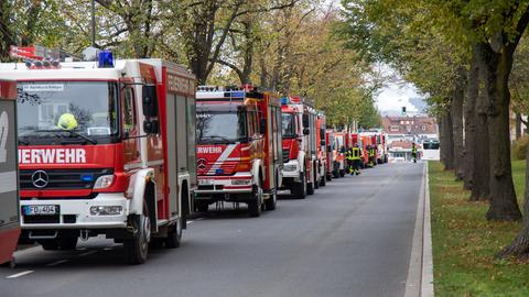 Das Bild zeigt eine breite, von Laubbäumen gesäumte Straße. Am linken Bildrand parken mehrere Feuerwehrautos hintereinander.