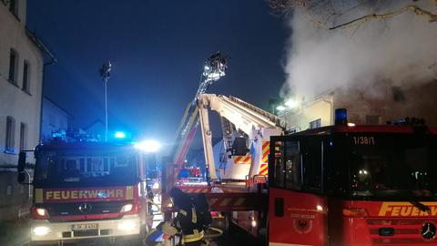 Dachstuhlbrand - Rauch kommt aus dem Dachstuhl eines zweigeschossigen Hauses - zu sehen im Hintergrund. Im Vordergrund ein Einsatzfahrzeug der Feuerwehr