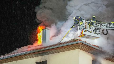 Wohnhausbrand in Büttelborn-Worfelden - aus dem Dach eines Wohnhauses schlagen Flammen - Feuerwehr mit Blaulicht und ausgefahrener Drehleiter löschen