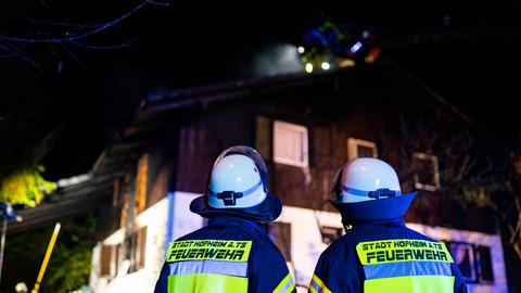 Im Vordergrund stehen in voller Montur zwei Feuerwehrmänner. Sie blicken nach oben auf einen immer noch rauchenden hölzernen Dachstuhl.