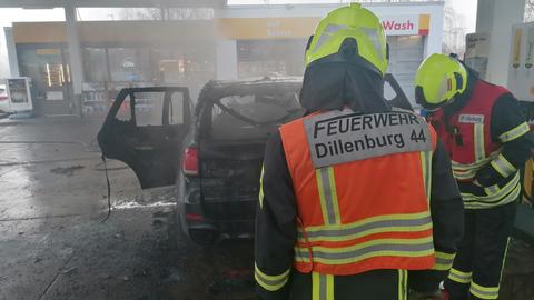 Ein Feuerwehrmann mit einer Jacke mit der Aufschrift "Feuerwehr Dillenburg" steht vor einem Auto. Das Auto ist ausgebrannt und qualmt vor Rauch.