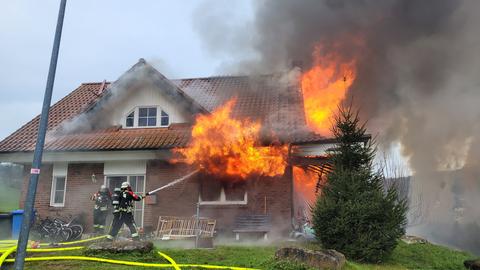 Zwei Feuerwehrleute löschen mit einem Schlauch aus dem Fenster eines Backstein-Einfamilienhauses schlagende Flammen.