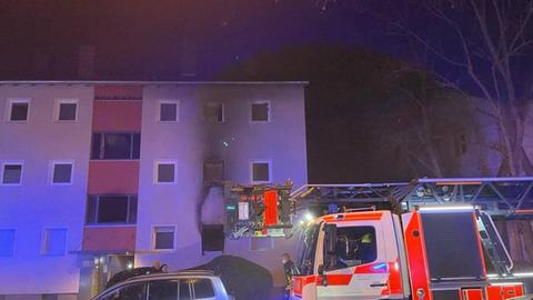 Das Bild zeigt ein Mehrfamilienhaus im Morgengrauen. Aus dem Fenster einer Erdgeschosswohnung kommt grauer Rauch. Vor dem Haus steht ein Feuerwehrauto mit ausgefahrener Rettungsleiter. Außerdem parken weitere Pkw vor dem Haus.