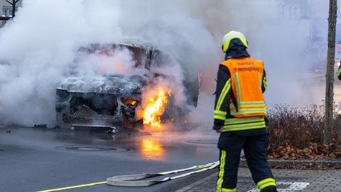 Ein brennendes Fahrzeug in einem Wohngebiet - im Vordergrund ein Feuerwehrmann im Einsatz. Das Auto brennt im vorderen Bereich, Rauch steigt auf. 