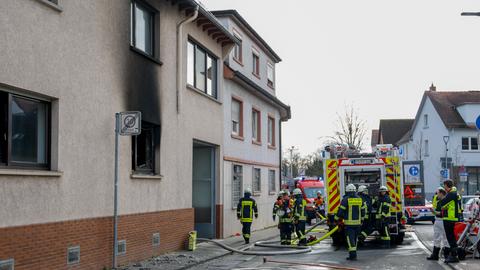 Fenster ohne Glasscheibe mit starken Rußspuren an einem Haus, Feuerwehrfahrzeug und Einsatzkräfte auf der Straße