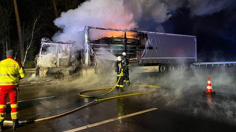 Feuerwehrmänner stehen vor einem ausgebrannten Lkw auf einer Straße und bespritzen ihn mit Wasser aus einem Schlauch. Rauch steigt in den nächtlichen Himmel.